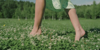Soft, barefoot steps through a clover field contrasted with a model wearing a simple gold chain, capturing the natural elegance that inspires Abellé’s limited-edition enamel cross necklaces.