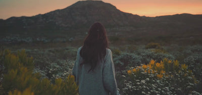 A woman walks through a field of wildflowers at sunset, capturing a peaceful, faith-filled moment that reflects the gentle beauty behind Abellé’s hand-painted cross necklaces.